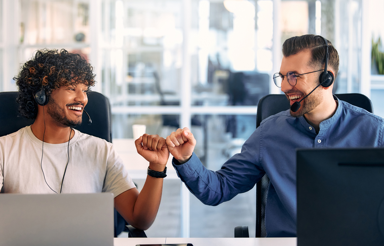 Two colleagues wearing headsets smiling and fist-bumping at desks in an office environment.