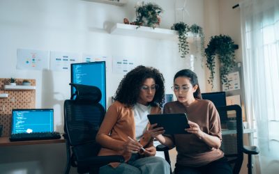 Two women collaborate on a tablet in a tech workspace, surrounded by code on computer screens.