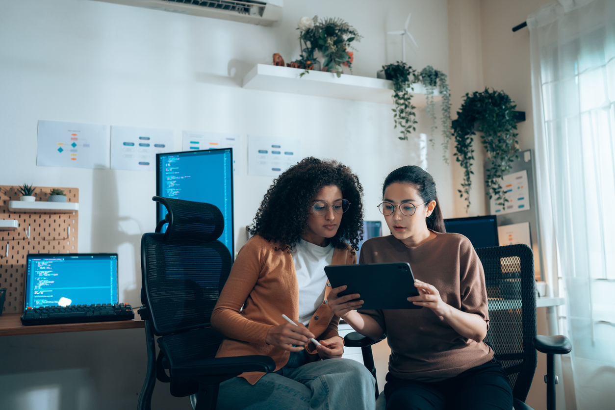 Two women collaborate on a tablet in a tech workspace, surrounded by code on computer screens.
