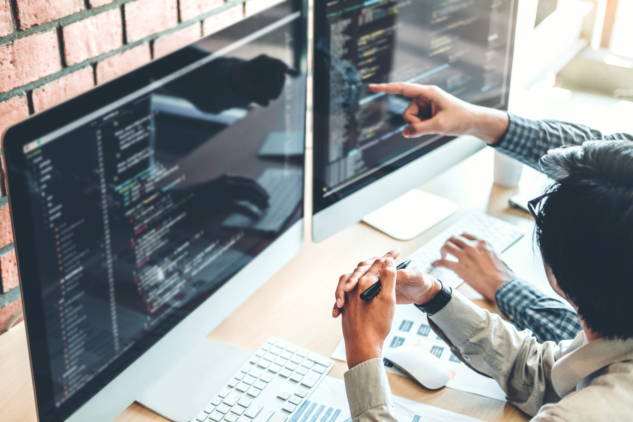 Two people collaborating on coding at dual computer monitors in a modern office setting.