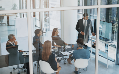Business meeting in a glass-walled office with a man presenting to a seated group of professionals.