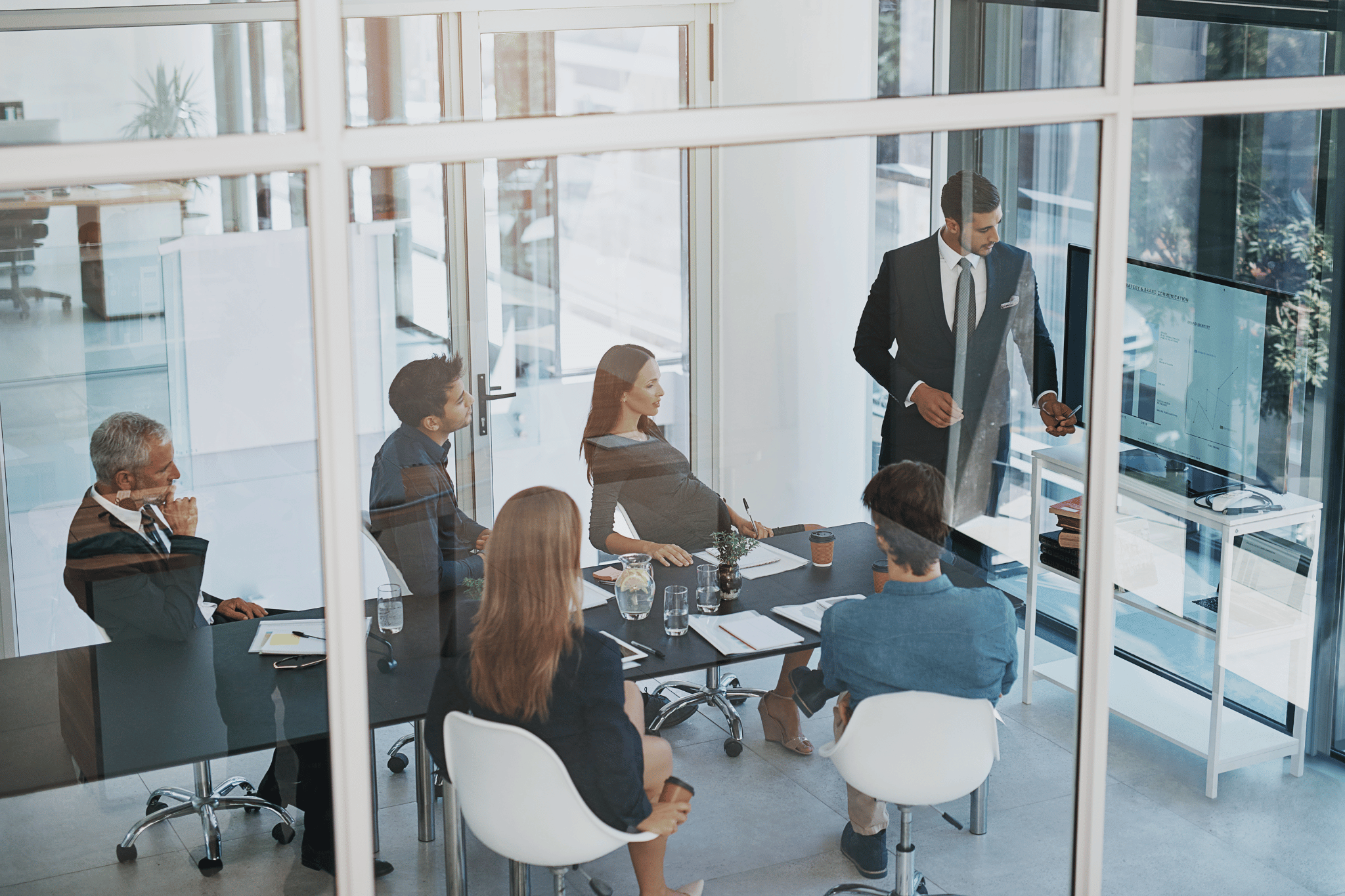 Business meeting in a glass-walled office with a man presenting to a seated group of professionals.